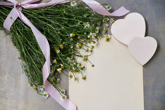 Small camomiles bouquet tied with pink ribbon and beige paper sheet on grey wooden table. Natural background picture of wild flowers. Ecological texture for poems, letters, romantic notes.