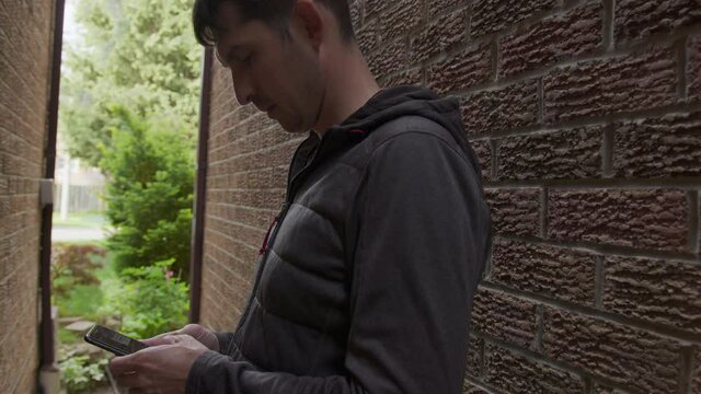 High quality 4k video of a young man who is texting messages on his cellphone standing in a shade of the red bricks wall. Guy hiding between the walls and holding mobile phone in his hands.