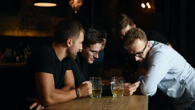 Five friends are relaxing in the pub. Guys watch videos on their smartphones and drink beer.