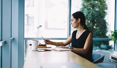 Prosperous attractive businesswoman dialing number from notepad making call to banking service, professional editor checking email box texting feedback using modern smartphone connected to wifi