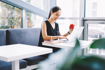 Positive formally dressed female journalist making distant job during work break in cafe interior typing messages in social networks communicating with followers,young entrepreneur satisfied with work