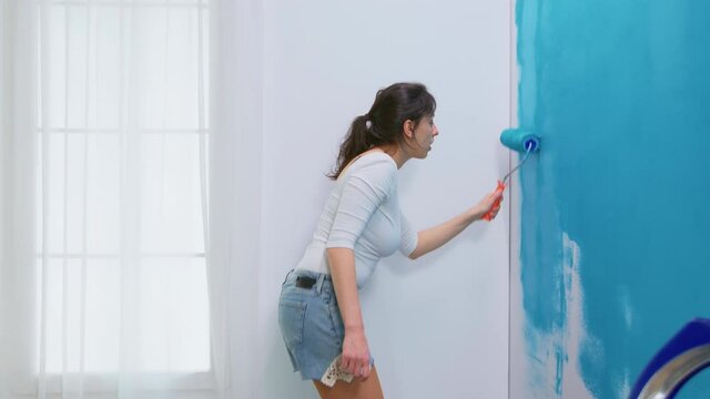 Woman decorates her home painting the wall with roller brush and dancing. Apartment redecoration and home construction while renovating and improving. Repair and decorating.