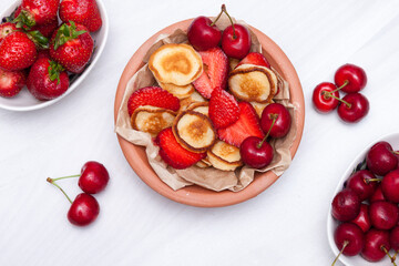 Mini tiny pancakes with straeberries and cherries on white wooden background. Trendy food concept. Flat lay.