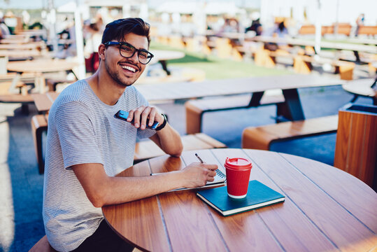 Portrait of happy bearded hipster guy planning working schedule sitting in cafe terrace in morning starting day with coffee,cheerful student excited with good news from university while doing homework - Powered by Adobe