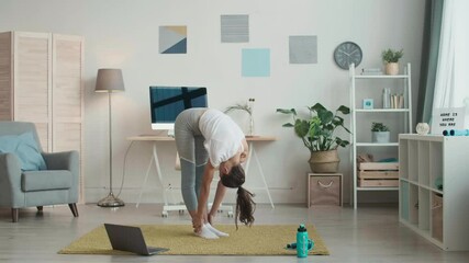 Wide shot of young Caucasian sporty woman wearing grey leggings and white t-shirt is standing on mat at laptop and doing stretching exercises