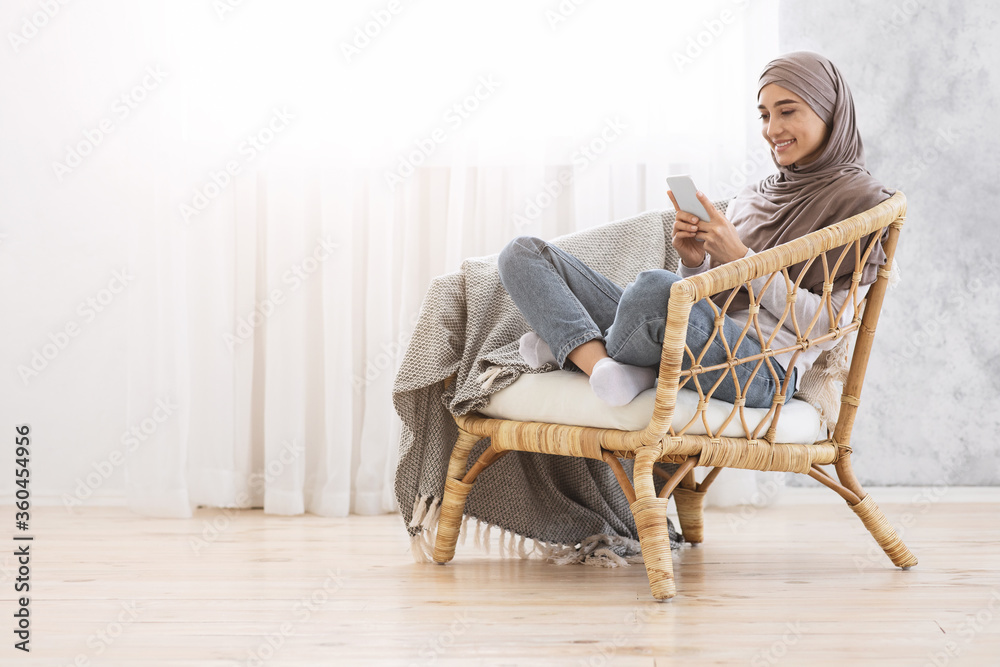 Wall mural smiling muslim woman sitting in modern chair with smartphone, relaxing at home