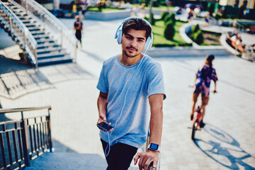 Pensive hipster guy in headphones walking on street with music using application on mobile for searching songs, young man strolling during sunny spring day enjoying playlist compositions via devices