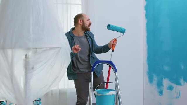 Repairman singing on roller brush dipped in blue paint while redecorating apartment. Housework, design, renovation. Home construction while renovating and improving. Repair and decorating.