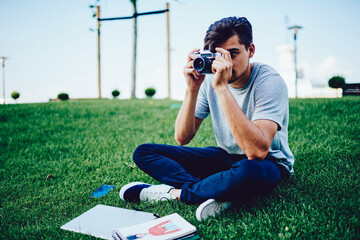 Young skilled photographer making photos on old vintage camera sitting on green lawn with stuff, hipster guy spending time on hobby concentrated on taking image on retro camera during sunny day