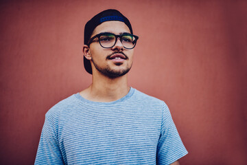 Handsome young male in casual t-shirt and black cap standing on wall promotional background,confident hipster guy in stylish spectacles and cool street look with mock-up for brand name or label