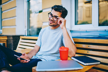 Cheerful hipster guy in stylish eyewear laughing while share media in social networks via 4G internet connection on smartphone resting on wooden bench ion street in urban setting enjoying leisure time