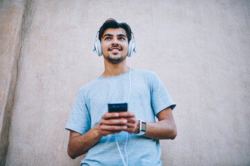 Below view of positive male meloman enjoying favourite playlist downloaded on smartphone via 4G...