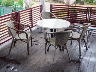 Wicker chairs stand around a white round table on a wooden floor wet after rain in an outdoor cafe