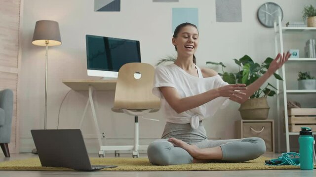 Wide shot of young female yoga tutor wearing grey leggings and white t-shirt is sitting on mat, looking at camera and telling about training purpose