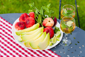 Summer party outdoor. Romantic picnic for a couple. Two glasses of cool wine served with mint and lime. Fresh melon and peach as simple light meal. Tasty low calories dessert. Green background.