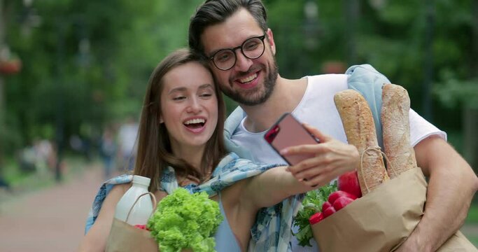 Joyfull couple smiling and posing for selfies with paper bags full of food. Good looking man and woman in 30s making photos on smartphone frontal camera while walking. Outdoors