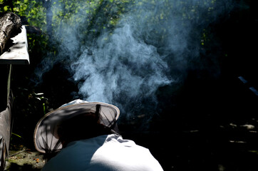 the beekeeper kneels in a white jumpsuit with a hood and a hat to the smoker who must light to burn...