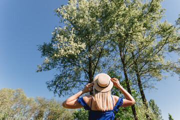 Portrait of a young woman wearing straw hat and blue dress standing back in rural landscape. Summer time, active life concept.