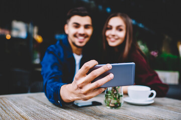 Selective focus on smartphone in hand of young man sitting with girlfriend on cafe terrace posing for selfie, cheerful male and female hipsters making picture for share in networks using mobile phone
