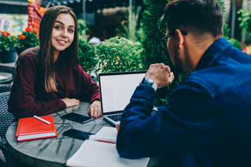 Portrait of cheerful teen brunette woman sitting at table on cafe terrace with male colleague using laptop computer with mock up screen for learning, back view of guy searching information on netbook