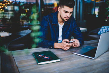 Thoughtful male student synchronizing mobile files with laptop for editing in application, pensive...