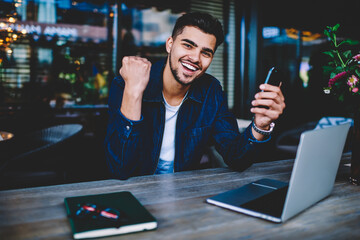 Cheerful hipster guy looking at camera excited with getting good news checking email on smartphone,...
