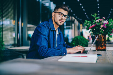 Portrait of serious caucasian male in spectacles working remotely on laptop computer, pensive hipster guy in spectacles for vision correction keyboarding publication for blog looking at camera