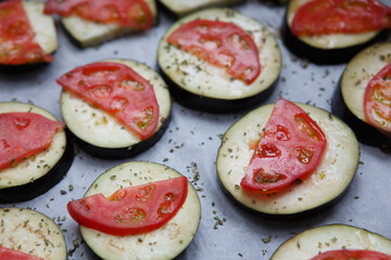 isolated filled frame close up shot of uncooked raw sliced aubergine eggplant with thin tomato slices on top sprinkled with dry green basil and oregano herbs, lying on a white baking paper background