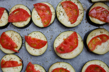 isolated filled frame close up shot of uncooked raw sliced aubergine eggplant with thin tomato slices on top sprinkled with dry green basil and oregano herbs, lying on a white baking paper background