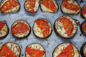 isolated filled frame close up shot of baked sliced aubergine eggplant with thin tomato slices on top, sprinkled with dry green basil and oregano herbs, lying on a white baking paper background