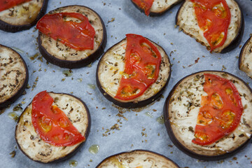 isolated filled frame close up shot of baked sliced aubergine eggplant with thin tomato slices on top, sprinkled with dry green basil and oregano herbs, lying on a white baking paper background