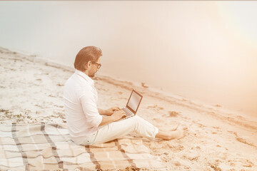Fototapeta na wymiar Copy writer works with laptop at sea side. Side view of smart caucasian businessman using computer while sitting on blanket near sea. Tinted image.
