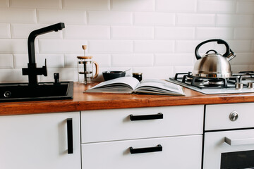 Big cook book on the wooden table. Metallic teapot on the gas oven. White kitchen interior. Wooden complete kitchen with gas oven.