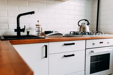 Big cook book on the wooden table. Metallic teapot on the gas oven. White kitchen interior. Wooden complete kitchen with gas oven.