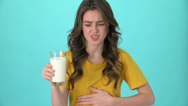 A displeased young woman wearing a yellow t-shirt has got lactose intolerance while holding a glass of milk standing isolated over blue background