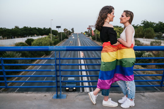 Two girls with a rainbow flag of LGBT pride on a bridge overlooking a highway with traffic