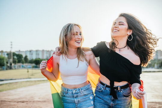 Two young girls (15-20 years old) walk around laughing with the LGBT Pride flag at sunset