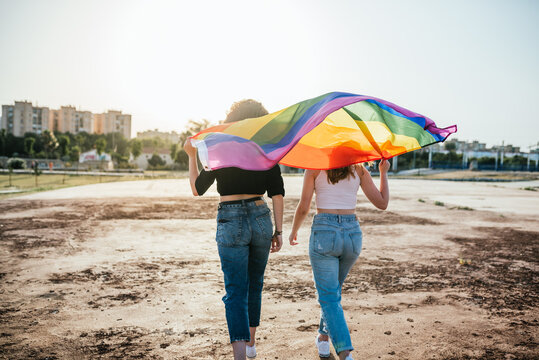 Two young girls (15-20 years old) on their backs walking with the rainbow flag of LGBT pride at sunset