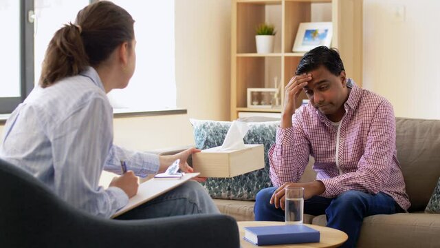 psychology, mental therapy and people concept - woman psychologist giving box with paper tissues to crying young indian male patient at psychotherapy session