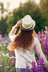 girl with her hair in a lupine field in a white lace blouse stands with her back to the camera