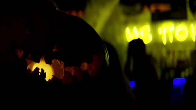 Halloween pumpkins in a spooky dark and lights. Stock footage. Close up of scary pampkin with a yellow lamp inside and people having fun on the background.