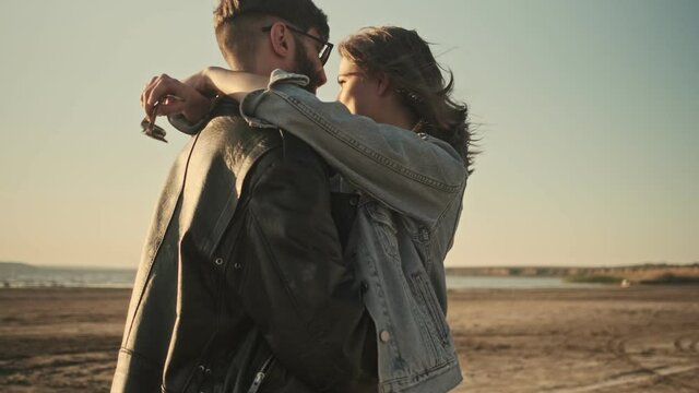 A side view of a smiling young couple man and woman are hugging and looking at each other while walking on the beach together during sunset