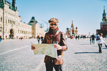 Obraz premium Young caucasian traveller dressed in trendy clothing searching location on paper map, stylish hipster guy trying find new sightseeing for discover while walking on Moscow Red Square during vacation