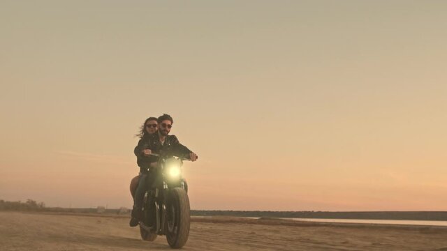 A happy young couple man and woman are riding on a motorbike together on the beach during sunset time with headlight on