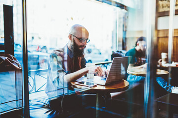 Millennial male blogger in eyeglasses searching information for content article on web page about software development, young freelancer working remotely in coffee shop watching videos on laptop