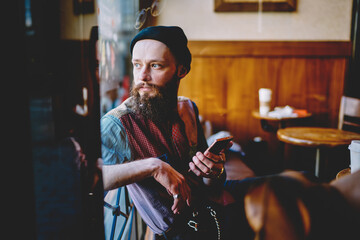 Thoughtful hipster guy pondering on creating publication and article for own web page, pensive bearded man holding smartphone gadget in hands and thinking about notification for mobile phone