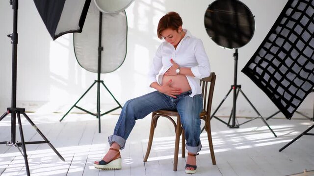 Pregnant woman sitting in a studio with reflector equipment full body view in white shirt massaging her belly. Young woman with short hair expecting a baby. Close up. Prores 422.