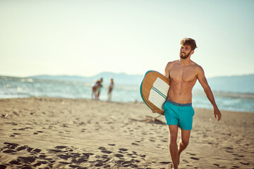 fit young beardy male walking on the sunny beach, holding surfboard, smiling. Copy space