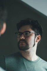 a handsome young man with a thick beard and glasses is illuminated on one side by a soft light. fashionable beard, happy. reflection in the mirror. irritation after shaving. shaves three times