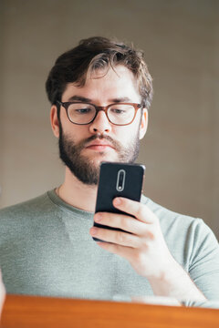 a handsome young man with a thick beard and glasses is holding a phone in his hands. take a photo. read the news. he looks at the phone carefully